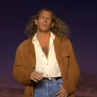 Michael Bolton can be seen wearing a brown jacket with denim jeans and a white shirt.